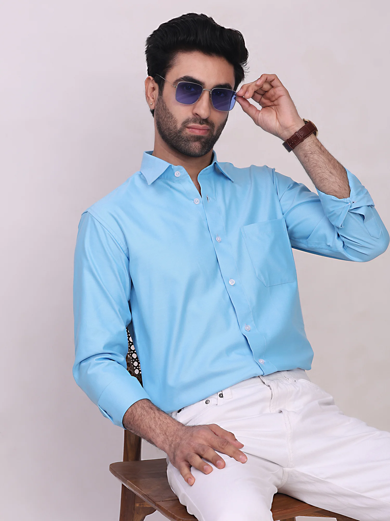 Front view of a model sitting, wearing a light blue long-sleeve shirt with a front pocket and white trousers