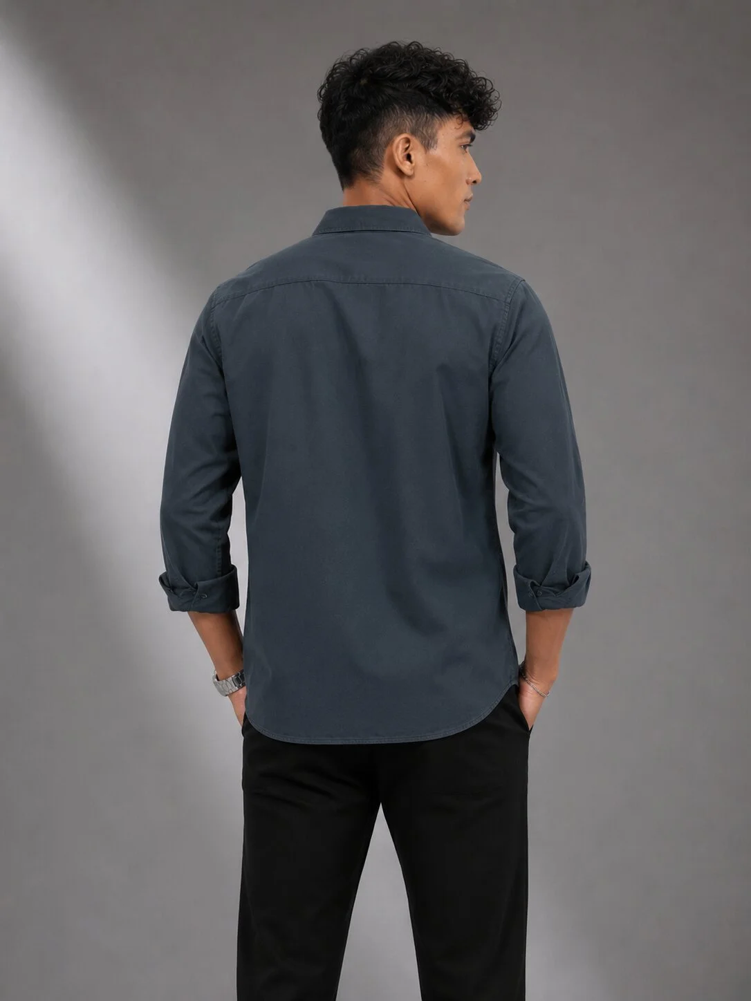 Back view of a male model wearing a charcoal grey long-sleeve shirt with rolled-up cuffs and black trousers